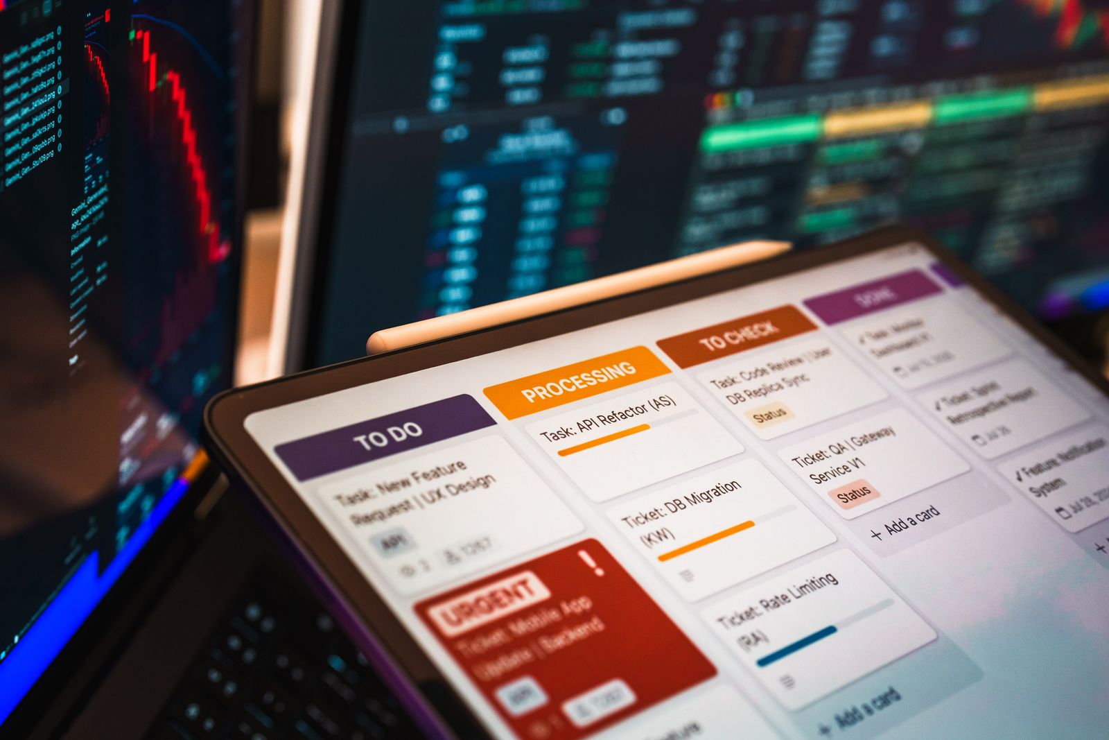 A tablet displaying a workflow task board, with data monitors in the background